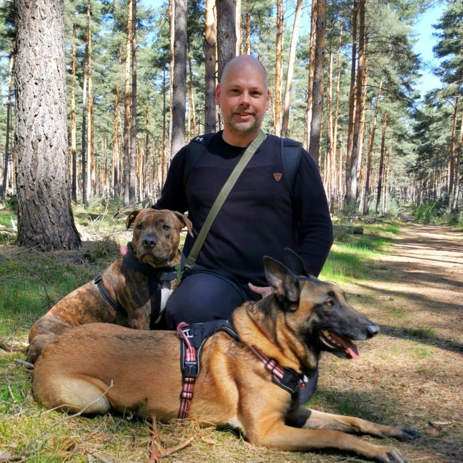Mann (Sven Mrosko) mit zwei Hunden im Wald, umgeben von hohen Bäumen und natürlichem Licht.