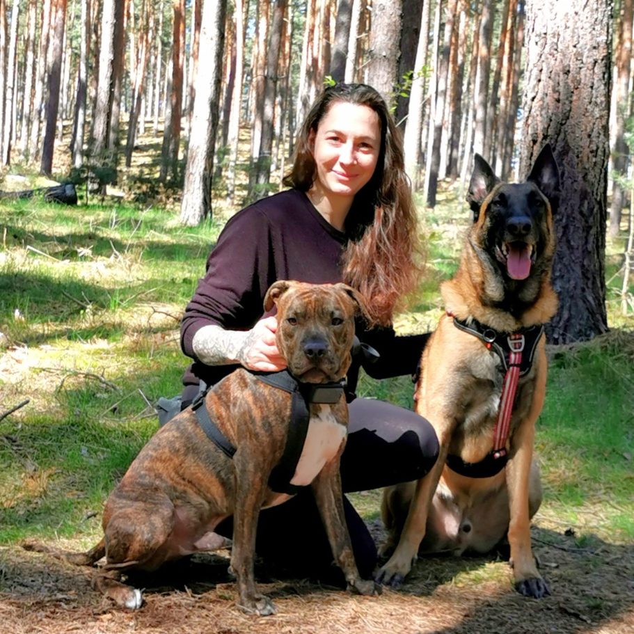 Frau mit zwei Hunden in einem Wald, umgeben von Bäumen und grünem Boden.